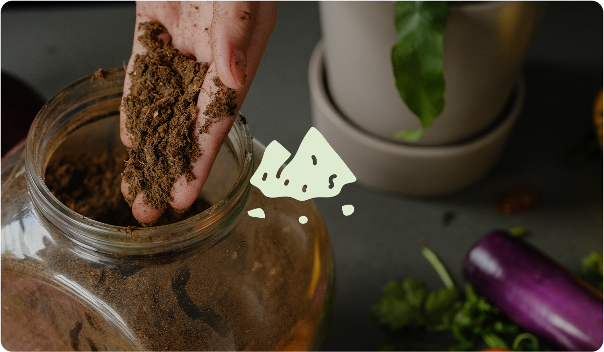 A person’s hand holds a handful of dry, dark brown organic fertilizer over a large glass jar. In the center is a simple green icon of a small mound of powder. Fresh vegetables and a potted plant are visible in the blurred background.