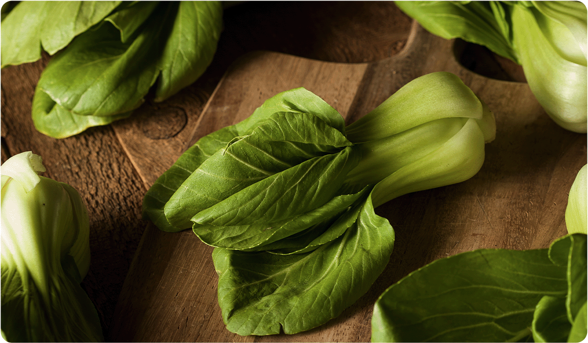 Several heads of fresh baby bok choy rest on a rustic wooden cutting board. The leaves are a vibrant green with thick, pale white stems, shown in warm, natural lighting.